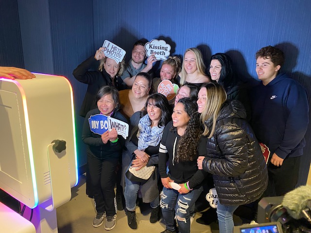 A group of people pose closely together in front of a photo booth, smiling and holding fun signs like “Kissing Booth” and “I Hit the Photo.” The background is a plain blue wall.