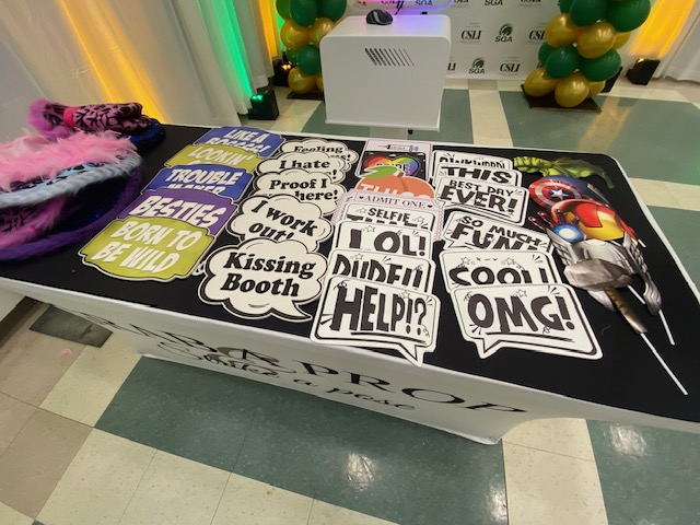 A table displays various colorful photo booth props and speech bubble signs with phrases like "OMG!", "Did it! Help?!", "Besties," and "Selfie," with balloons and lights in the background.