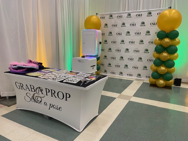 A photo booth setup with a table holding props, a photo booth machine, and a backdrop with the CSU and SGA logos. Large green and gold balloon columns stand to the right of the backdrop.