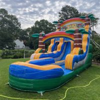 A large, colorful inflatable water slide with palm tree decorations stands on a grassy lawn, surrounded by greenery and trees, with a house visible in the background under a partly cloudy sky.