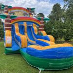 A large, colorful inflatable water slide with palm tree decorations is set up on a grassy lawn, surrounded by trees and greenery. The slide features two lanes and a curved splash pool at the bottom.
