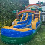 An inflatable water slide with palm tree decorations and bright blue, yellow, and green colors is set up on a grassy lawn next to a house and tall bushes.