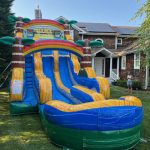 A large, colorful inflatable water slide with palm tree decorations is set up on a grassy lawn in front of a house. A person stands nearby, and the sky is clear and sunny.
