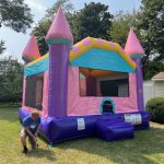 A person stakes down a large, colorful inflatable bounce house shaped like a castle, set up on grass in a backyard with trees and a small white shed nearby.