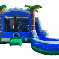A blue and green inflatable bounce house with palm tree decorations and an attached curved slide, isolated on a white background.