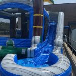 An inflatable water slide with a palm tree design, blue slide, and splash pool at the bottom, set up outdoors near a building and metal railings.