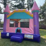 A colorful inflatable bounce house shaped like a castle with pink, purple, and yellow accents sits on a grassy lawn with trees and a fence in the background.