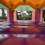 Interior view of an empty, colorful inflatable bounce house with orange, purple, and pink sections, taken from the entrance, showing tree branches and greenery visible through the mesh windows.