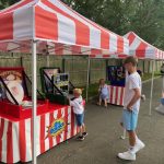 Children and adults play carnival games under red-and-white striped tents set up outdoors on a paved surface, with trees and a wooden fence in the background.