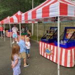 Children and adults enjoy a carnival with red and white striped tents and various game booths set up outdoors on a paved area near a wooden fence and trees.