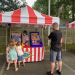 Two children and an adult stand in front of a carnival game booth with red and white striped tents. The booth has colorful game boards, and a staff member stands behind the counter. Trees and a wooden fence are in the background.
