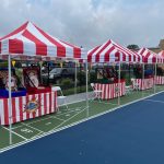 Red and white striped tents are set up in a row on an outdoor court, each with carnival-style games and prize tables underneath. White folding chairs are placed beside each game booth. The sky is partly cloudy.