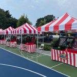 Red-and-white striped tents are set up in a row on an outdoor court, each with a carnival game inside. Folded chairs sit in front of the booths, and trees and parked cars are visible in the background.