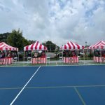 Four red-and-white striped tents are set up in a row on a blue outdoor court, each with a table underneath displaying carnival-style games or decorations. Cars and greenery are visible in the background under a cloudy sky.