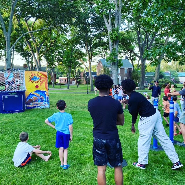 A group of children and adults gather on green grass in a park, watching a dunk tank game. Some kids are sitting or standing, while a man on the dunk tank platform waits to be dunked. Large trees and buildings appear in the background.