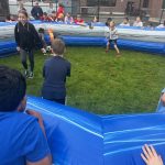 Children play a game with a soccer ball inside an inflatable blue and gray ring, while other kids watch from outside the barrier. The event takes place outdoors on grass near a brick building.