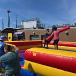 Two people stand on an inflatable jousting arena, each holding a padded stick, while others watch and staff supervise outside on a sunny day near a sports facility.