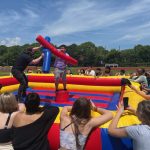 Two people stand on an inflatable jousting arena, each holding large foam batons, while a crowd of spectators watches and cheers outdoors under a sunny sky.