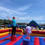 A person stands with arms raised on a red podium in the middle of a colorful inflatable obstacle course, while people gather around outside on a sunny day.