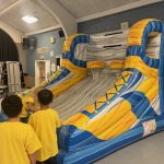 Two children in yellow shirts stand in front of a large inflatable basketball game shaped like sneakers inside a gymnasium. The game features hoops and basketball lanes with blue and yellow designs.