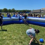 Children play a ball game inside an inflatable blue and white arena on a grassy field, while a crowd of spectators watches. A school building and trees are visible in the background under a sunny sky.