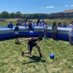 Children play a game with a blue ball inside an inflatable circular arena on a grassy field, while others watch from the outside. The day is sunny, and a school building is visible in the background.