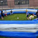 Several children play soccer inside an inflatable blue and gray arena on grass, with a brick building and fence in the background. The kids are actively chasing the ball.
