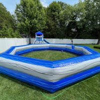 An inflatable blue and gray octagonal arena is set up on a grassy lawn, with a playset and slide visible in the background. The area is surrounded by white fencing and trees.