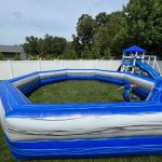 An inflatable blue and gray octagonal arena set up on grass in a backyard; a child inside runs while another inflatable slide structure is visible in the background.
