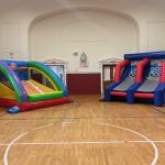 Two inflatable slides—one colorful with a climbing wall, the other blue and red with two lanes—are set up inside a gymnasium with wooden floors and red walls.