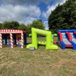 Three colorful inflatable game structures are set up on grass outdoors under a partly cloudy sky, with trees in the background. The inflatables feature red, white, green, and blue designs.