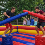 Two young people stand on round pedestals in an inflatable arena, jousting with large padded batons. A group of onlookers cheer enthusiastically in the background. The scene takes place outdoors with trees behind them.