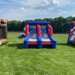 Three inflatable carnival games are set up on a grassy field, including an axe-throwing game, a basketball shooting game, and a football toss. Trees and a clear sky are visible in the background.