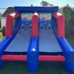 An inflatable skee-ball game set up outdoors on grass, featuring two blue and red lanes with targets labeled "Roller Ball" at the back and a few green balls on the ground.
