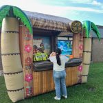 A person in a white shirt and jeans stands at an inflatable tiki bar decorated with palm trees and colorful designs. A sign reads "Open Tiki Bar." The setup is outdoors near a brick building on a sunny day.