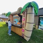 A person sets up supplies at an inflatable tiki bar decorated with palm trees and flowers, outdoors on grass near a building. Boxes and drinks are visible on the counter.