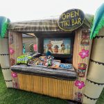 Inflatable tiki bar decorated with tropical designs and a sign reading "Open Tiki Bar." The counter displays various snacks and treats, and there’s a vibrant island scene backdrop inside the bar. The setup is outdoors on grass.