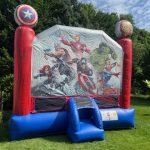 A large inflatable bounce house with Marvel Avengers characters depicted on the front, including Iron Man, Captain America, and Thor, stands on a grassy lawn under a partly cloudy sky.