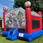 A red and blue inflatable bounce house featuring Marvel Avengers characters, including Iron Man, Captain America, Hulk, and Thor, with character graphics and large shield decorations on top. The bounce house is set up on grass.