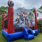 A colorful Avengers-themed bounce house sits on grass, featuring images of superheroes like Iron Man, Captain America, Thor, Hulk, and Black Widow on the front, with red, blue, and black accents.