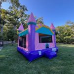 A colorful inflatable bounce house shaped like a castle, with pink and purple turrets, sits on a grassy lawn surrounded by trees on a sunny day. A car is parked nearby.