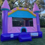 A colorful inflatable bounce house shaped like a castle with pink, purple, and yellow accents sits on a grassy lawn, surrounded by trees and a parked car in the background.