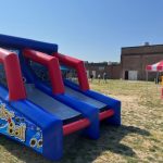 An inflatable blue and red "Roller Ball" arcade game sits on a grassy field near a red-and-white striped tent. A few people are visible in the background near a brick building on a sunny day.