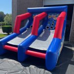 An inflatable blue and red basketball arcade game with two lanes is set up outdoors in front of a building, featuring scoring hoops and game graphics on the backboard.