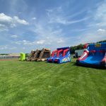Four colorful inflatable structures, including slides and an obstacle course, are set up on a large grassy field under a mostly sunny sky with some clouds. Trees and a building are visible in the background.