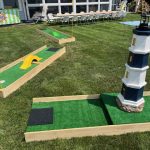A backyard mini golf setup on green grass features several putting greens, including one with a yellow ramp and another with a large lighthouse decoration. Tables and chairs are arranged near a house in the background.