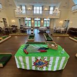 Mini golf course setup indoors with several putting greens and obstacles arranged around a large room. In the foreground, a table with putters and golf balls has a green and white striped cloth labeled "Big Bounce.