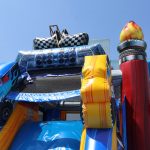 A colorful inflatable slide with a racing and car theme, featuring large tire and flame decorations, a checkered flag, and a blue monster truck, set outdoors on a sunny day.