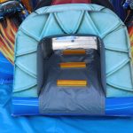 A close-up of the entrance to an inflatable obstacle course, featuring three yellow steps inside and a blue and gray archway, with a blue mat on the ground and racing-themed decorations in the background.