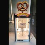 A glass display case for SuperPretzel soft pretzels with a large pretzel sign on top, empty shelves inside, and a power cord hanging out the side, placed on a white counter against a gray wall.